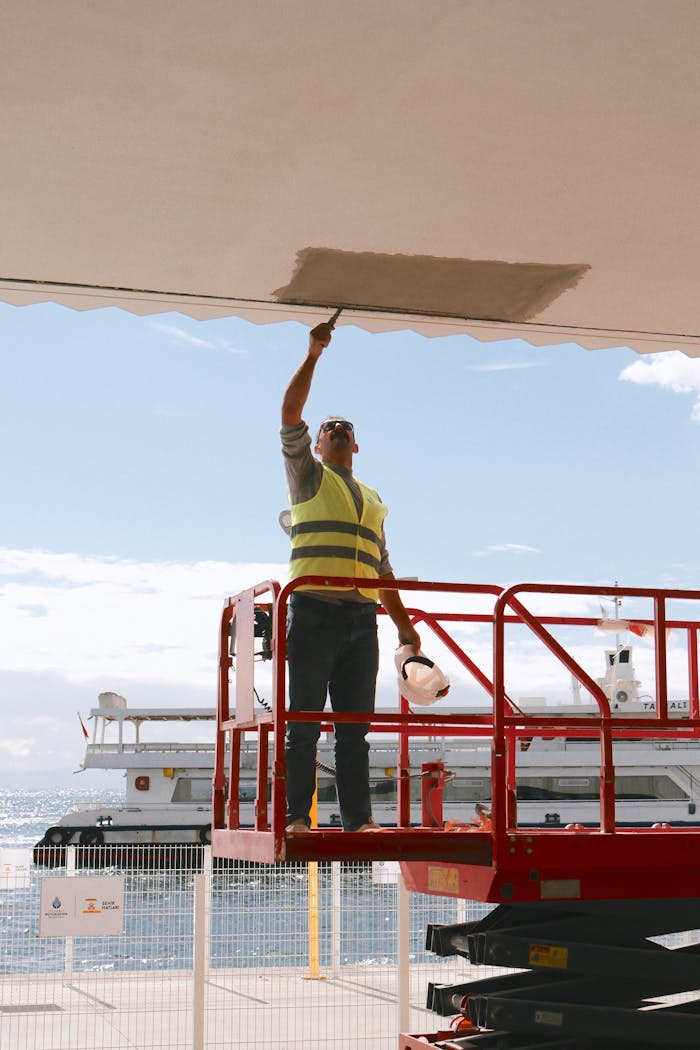 A construction worker in a safety vest paints a ceiling while on a lift by the sea, with a boat in the background.
